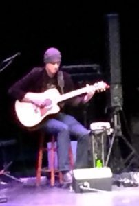 A man sitting on top of a chair playing an acoustic guitar.