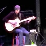 A man sitting on top of a chair playing an acoustic guitar.