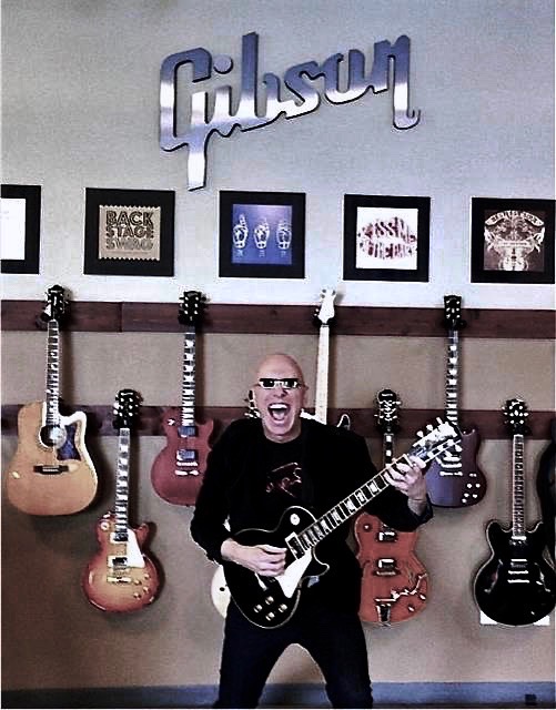 A man holding a guitar in front of many guitars.